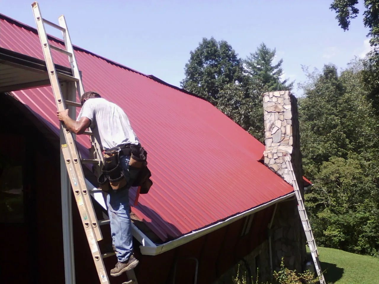 Roofer on a ladder beside a red metal roof and stone chimney — Soffit & Fascia Repair by Roofing Brothers in Enville